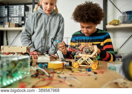 Young Engineer Using Soldering Iron To Join Chips And Wires. His Friend Looking At The Process. Robo