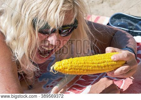 The Blonde With Her Mouth Wide Open Wants To Bite A Large Ear Of Boiled Corn With Her Teeth. Close-u