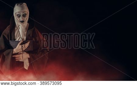 An evil cursed nun with a bible and a cross in her hands looks at the camera on a black background with a blood-red haze. Halloween. Horrors. Copy space.