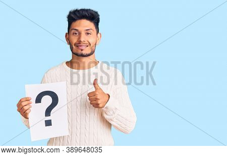 Handsome latin american young man holding question mark smiling happy and positive, thumb up doing excellent and approval sign 