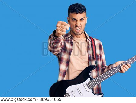 Young hispanic man playing electric guitar annoyed and frustrated shouting with anger, yelling crazy with anger and hand raised 