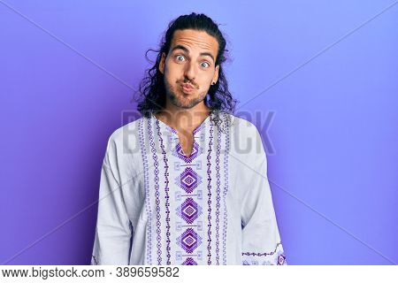Young handsome man with long hair wearing bohemian and hippie shirt puffing cheeks with funny face. mouth inflated with air, crazy expression. 