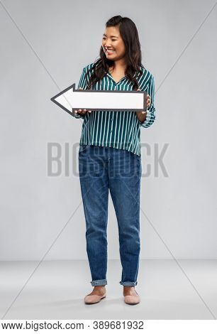 direction and people concept - happy smiling asian young woman holding big white leftward thick arrow over grey background
