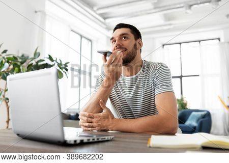 technology, remote job and people concept - happy smiling man with laptop computer calling on smartphone or using voice command recorder at home office