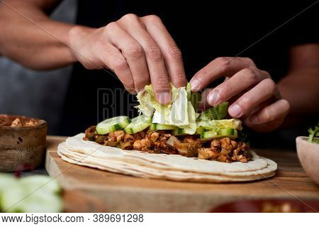 closeup of a young caucasian man preparing a durum or a burrito, with chicken meat cooked with different vegetables such as onion or red and green pepper, and fresh lettuce and raw cucumber