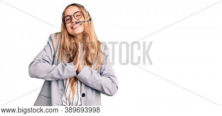 Young beautiful blonde woman wearing call center agent headset praying with hands together asking for forgiveness smiling confident. 