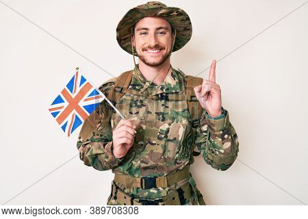 Young caucasian man wearing camouflage army uniform holding united kingdom flag surprised with an idea or question pointing finger with happy face, number one 