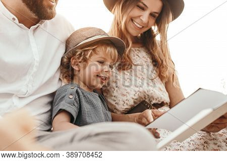 Low Angle Of Delighted Boy Reading Fairytale With Crop Mother And Father During Picnic In Countrysid