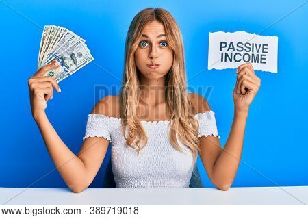 Beautiful blonde young woman holding dollars and passive income text puffing cheeks with funny face. mouth inflated with air, catching air. 