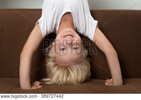 Cheerful Fair-haired Boy Stands In His Arms And Smiles. Physical Education At Home.