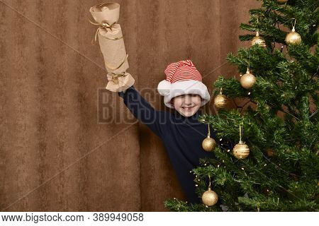 A Boy With A Homemade Flapper, Peeps Out From Behind The Christmas Tree. He Smiles Cheerfully.