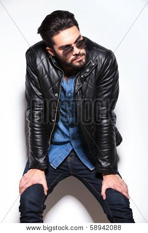 young man in leather jacket and sunglasses looking to his side while resting against studio wall