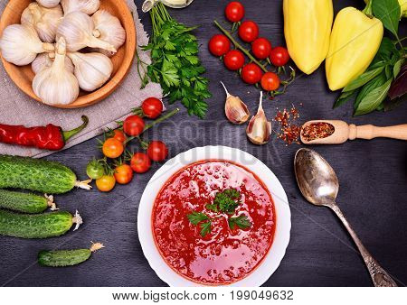 Fresh cold tomato and vegetable soup gazpacho in a white round plate with an iron spoon on a black background around the ingredients and spices