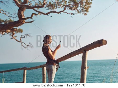 A woman stands near a wooden fence on the sea beach and takes pictures on a smartphone