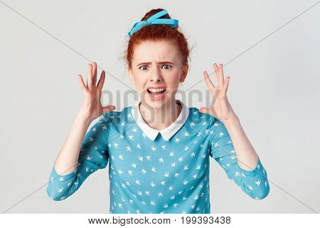 girl in despair and shock. Portrait of young angry redhead girl in blue dress looking panic screaming in camera with mouth wide open with hands up. Isolated studio shot on gray background.