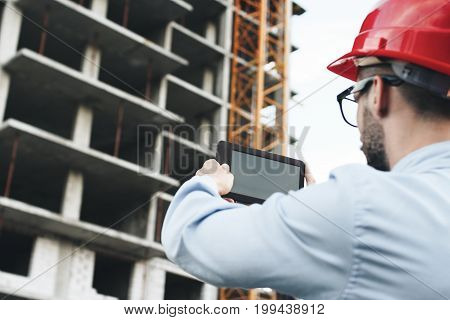 Young businessman or engineer holding modern tablet and scans building on construction. Portrait of engineer in red hard hat with gadget on construction site
