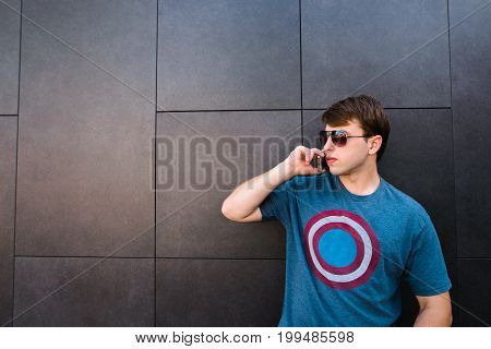 Young man in glasses talking on a mobile phone and looking side by side against a gray wall.