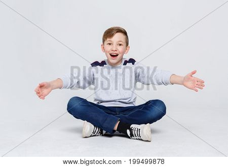 Ready to hug the world. Happy boy in braces sitting legs crossed on studio floor. Kid in casual laughing, posing on camera at white background, copy space