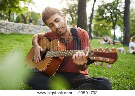 Outdoor portrait of talanted man playing musical instrument and enjoying summer day. Bearded musician playing outdoors, sitting cross-legged on a grass.