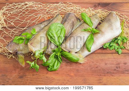 Several uncooked carcasses of the notothenia fish without of head and tail and twigs of basil and parsley on the fishing net on a dark wooden surface