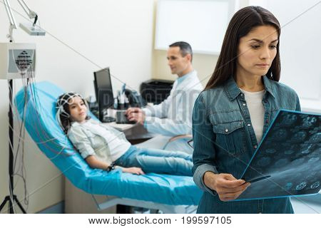 Worrying results. Charming young mother holding the results of computer tomography and carefully examining them, looking worried, while her little daughter undergoing electroencephalography