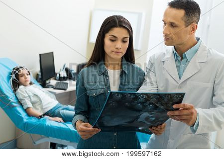 Nothing to worry about. Pleasant middle-aged doctor showing the results of computer tomography to the mother of a little girl undergoing electroencephalography