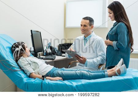Laid-back conversation. Charming young doctor putting pleasantly talking to a little girl undergoing encephalography and putting down her answers while her mother standing next to him
