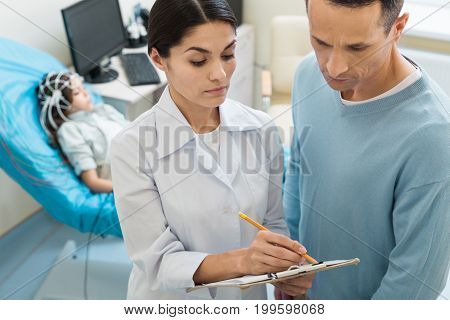 Necessary formalities. Beautiful female doctor with a sheetholder asking the father of a little girl undergoing electroencephalography to sign the documents
