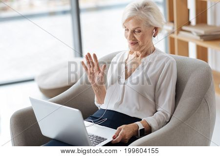 Nice to see you. Joyful positive senior woman wearing headphones and looking at the laptop screen while greeting her friend