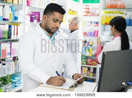 Chemist Writing On Clipboard While Colleagues Working In Pharmac