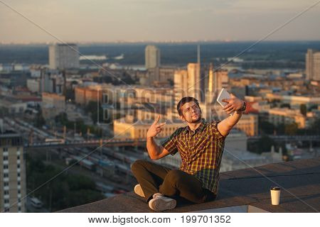 A hipster guy makes a selfie on the roof of a high-rise building. The blogger has conquered a new height. The video blogger broadcasts live from the roof.