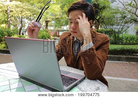 Tired young business man holding glasses and suffering eyestrain in front of screen laptop at outdoor park.