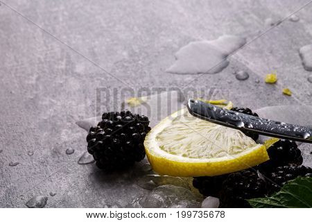 A gray table with sappy blackberries, drops of water, yellow slice of lemon, fresh green leaves of mint and a metallic moist spoon on a gray blurred background.