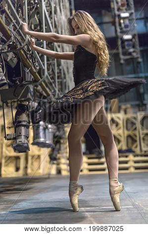 Gorgeous ballerina stands sideways on pointes next to the big stage light grip in the interior. She wears a black tutu with colorful rhinestones. Girl holds her hands on the grip and looks downward.
