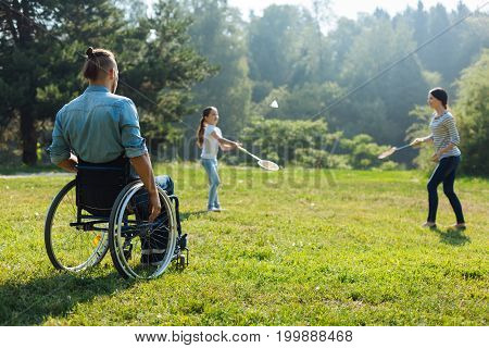 Enjoying sports. Pleasant young man with mobility impairment sitting in the wheelchair and watching his family play badminton in the meadow