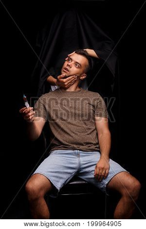A deeply stressed student sitting on a chair holding a drug syringe on a black background. The Death in black clothing holding a head of a narcomaniac man. Helpless male dying from drugs overdose.