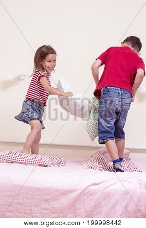 Kids Concepts and Lifestyle. Caucasian Brother and Sister Having Funny Pillow Battle on Bed Indoors. Vertical Image Composition