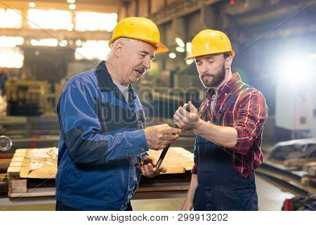 Two confident machinebuilding experts in workwear consulting about usage or features of workpiece in their hands
