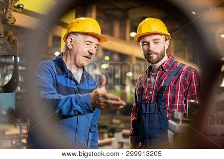 Aged master and his trainee in workwear and protective helmets consulting about new industrial machine equipment in workshop