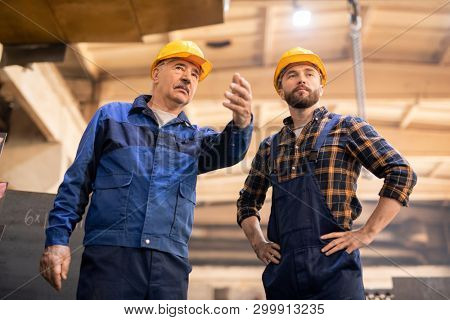 Young serious trainee looking at machine shown by his master while working together inside heavy industry plant