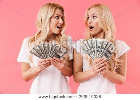 Shocked blonde twins wearing in t-shirts holding money and looking at each other oer pink background
