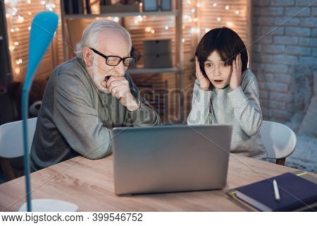 Grandfather And Grandson Are Watching Video At Laptop At Night. Grandfather Is Looking At His Grands