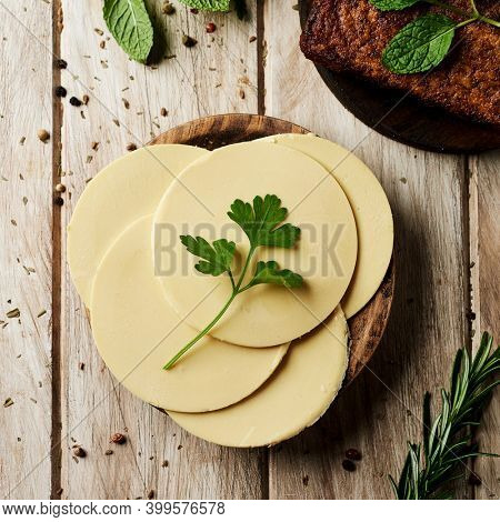 high angle view of some round slices of vegan cheese on a wooden plate placed on a white rustic table, next to some cooked tofu fillets