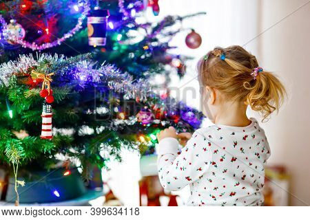 Adorable Toddler Girl In Pajamas Decorating Christmas Tree With Toy In Cute Hands. Little Child In N