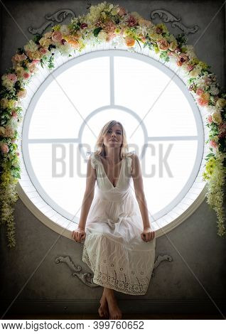 A Woman In A White Dress 35-45years Old Sits In A Round Window. She Looks Away.
