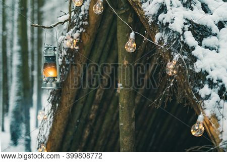 Kerosene Lamp And Garlands Hanging On Survival Shelter In Winter Forest