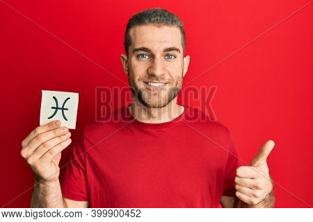 Young caucasian man holding paper with pisces zodiac sign smiling happy and positive, thumb up doing excellent and approval sign 