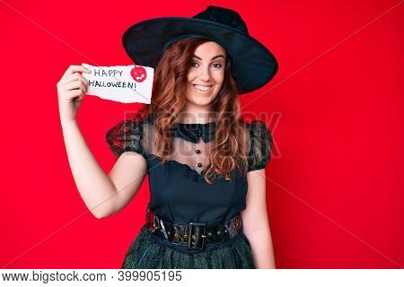 Young beautiful woman wearing witch costume holding happy halloween message paper looking positive and happy standing and smiling with a confident smile showing teeth 