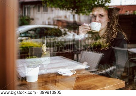 Cute Brunette, Curly Girl Sitting In A Cafe, Drinking Coffee And Juice. View Though The Wondow
