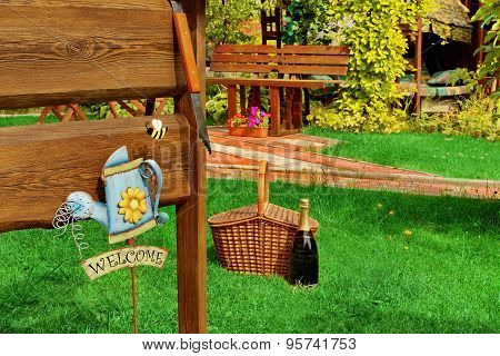 Outdoor Backyard Bbq Grill Party Or Picnic Scene In The Summer Outdoor Wooden Furniture Picnic Hamper Basket Champagne Wine Bottle Bbq Grill Tools Empty Wood Signboard Sign Garden Plants Trees In The Background